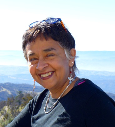 This is a headshot style photograph of Dr. Devaki Bhaya smiling at the camera with glasses on her head. Wearing silver jewelry and a black shirt, she is standing in front of forest covered mountains.